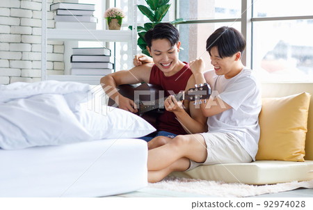 Two Asian young happy romantic lovely teenager male gay men lover couple partner sitting smiling on cozy sofa in bedroom cuddling hugging together while husband playing acoustic guitar and sing song Two Asian young happy romantic lovely teenager male gay men lover couple partner sitting smiling on cozy sofa in bedroom cuddling hugging together while husband playing acoustic guitar and sing song 92974028
