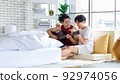 Two Asian young happy romantic lovely teenager male gay men lover couple partner sitting smiling on cozy sofa in bedroom cuddling hugging together while husband playing acoustic guitar and sing song 92974056
