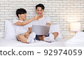 Two Asian romantic lovely teenager male gay men lover couple partner sitting smiling on bed in bedroom holding orange juice glass cuddling together watching streaming movie online via laptop computer 92974059