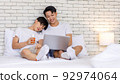 Two Asian romantic lovely teenager male gay men lover couple partner sitting smiling on bed in bedroom holding orange juice glass cuddling together watching streaming movie online via laptop computer 92974064