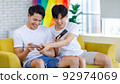 Two Asian cheerful teenager male gay men lover couple partner sitting smiling on sofa holding smartphone playing fun digital game online together in living room with rainbow pride equality unity flag 92974069