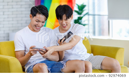 Two Asian cheerful teenager male gay men lover couple partner sitting smiling on sofa holding smartphone playing fun digital game online together in living room with rainbow pride equality unity flag Two Asian cheerful teenager male gay men lover couple partner sitting smiling on sofa holding smartphone playing fun digital game online together in living room with rainbow pride equality unity flag 92974069