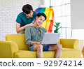 Asian teenager male gay businessman sitting on sofa holding laptop notebook computer working online from home while boyfriend help massaging shoulder in living room with rainbow equality pride flag 92974214