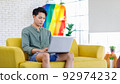 Asian young teenager male gay businessman sitting on sofa holding laptop notebook computer working online from home in living room with rainbow equality unity flag celebrate LGBTQ June pride month 92974232