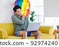 Asian young teenager male gay businessman sitting on sofa holding laptop notebook computer working online from home in living room with rainbow equality unity flag celebrate LGBTQ June pride month 92974240