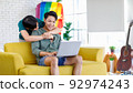 Asian teenager male gay businessman sitting on sofa holding laptop notebook computer working online from home while boyfriend help massaging shoulder in living room with rainbow equality pride flag 92974243