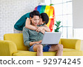 Asian teenager male gay businessman sitting on sofa holding laptop notebook computer working online from home while boyfriend help massaging shoulder in living room with rainbow equality pride flag 92974245