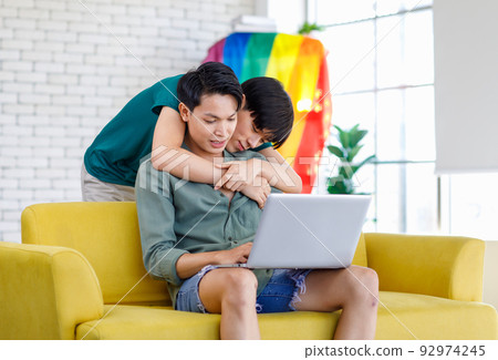 Asian teenager male gay businessman sitting on sofa holding laptop notebook computer working online from home while boyfriend help massaging shoulder in living room with rainbow equality pride flag Asian teenager male gay businessman sitting on sofa holding laptop notebook computer working online from home while boyfriend help massaging shoulder in living room with rainbow equality pride flag 92974245