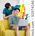 Asian teenager male gay businessman sitting on sofa holding laptop notebook computer working online from home while boyfriend help massaging shoulder in living room with rainbow equality pride flag 92974246