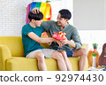 Asian happy teenager male gay man boyfriend sitting smiling on sofa giving anniversary wrapped present box to lover cuddling hugging together in living room with rainbow pride celebrate equality flag 92974430