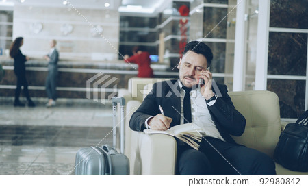 Pan shot of confident businessman talking mobile phone and writing notes in notepad while sitting on armchair in luxury hotel with luggage near him. Travel, business and people concept 92980842