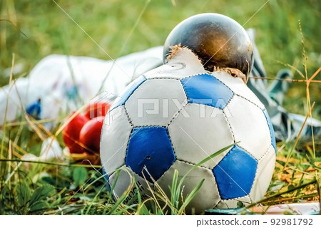 Burst soccer ball lies in pile of garbage on green grass in city. Torn ball thrown as rubbish after playing football game close view Burst soccer ball lies in pile of garbage on green grass in city. Torn ball thrown as rubbish after playing football game close view 92981792