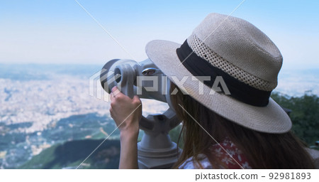 Unrecognizable woman traveler looking through viewing binoculars at summer city. Copy space. Woman in straw hat observes of blur panorama, with binoculars at observation deck. Mountain Dajti Albania 92981893