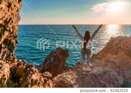 A female tourist, standing with her hands up, reaches the ends of the earth and sees the dragon rock. The girl welcomes the sun. Concept design. A successful woman looks at the sunset with open arms. A female tourist, standing with her hands up, reaches the ends of the earth and sees the dragon rock. The girl welcomes the sun. Concept design. A successful woman looks at the sunset with open arms. 92982028
