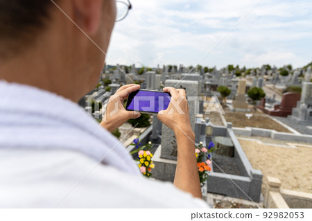 A senior man visiting a grave and taking a picture of the grave with a smartphone after cleaning 92982053