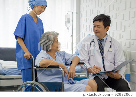 Young nurse push a wheelchair of the elderly patient while asian doctor with stethoscope and gown examine the symptom of the Total Knee Arthroplasty. Healthcare and medicine concept. 92982063