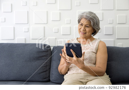 Asian gray hair old woman looking at tablet computer monitor, smiling while sit on sofa in the living room. Asian gray hair old woman looking at tablet computer monitor, smiling while sit on sofa in the living room. 92982176