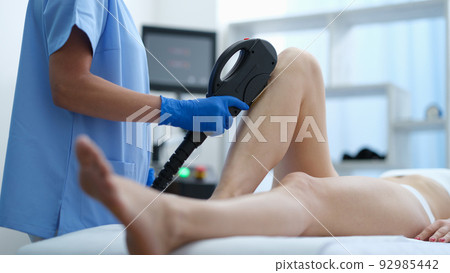 Hands of beautician in blue medical gloves hold depilator and use it on female legs Hands of beautician in blue medical gloves hold depilator and use it on female legs 92985442