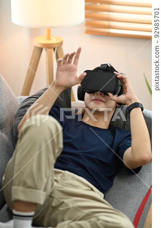 Man in casual clothes watching movie or play video game on virtual reality headset, lying on couch at home Man in casual clothes watching movie or play video game on virtual reality headset, lying on couch at home 92985701