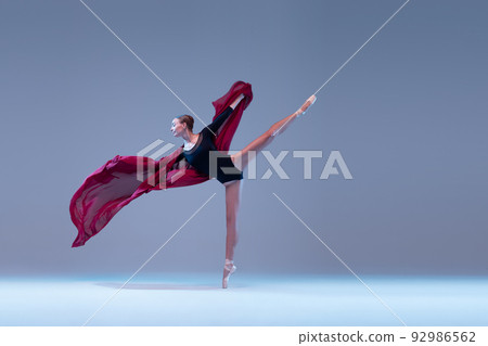 Portrait of young ballerina dancing with deep red fabric isolated over blue grey studio background. Standing on tiptoe 92986562