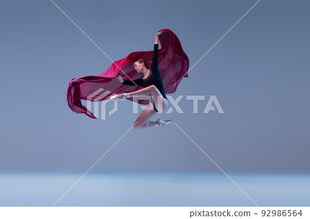 Portrait of young ballerina dancing with deep red fabric isolated over blue grey studio background. Graceful performance 92986564