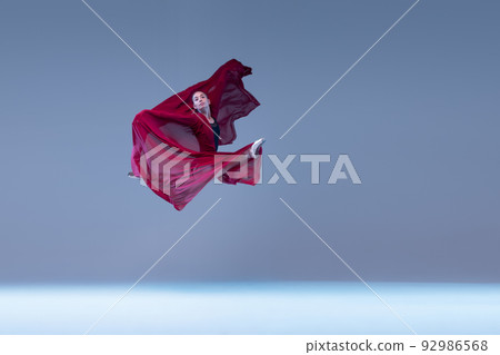 Portrait of young ballerina dancing covered with deep red transparent fabric isolated over blue grey studio background. Flying high 92986568