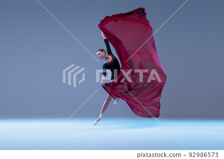 Portrait of young tender ballerina dancing with deep red fabric isolated over blue grey studio background. Lightness, grace 92986573