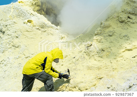 male volcanologist on the slope of a volcano next to a smoking sulfur fumarole examines a sample of a mineral male volcanologist on the slope of a volcano next to a smoking sulfur fumarole examines a sample of a mineral 92987367