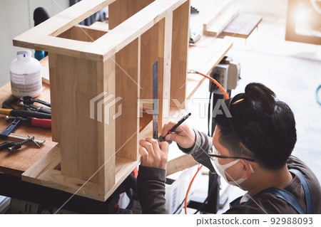A carpenter measures the planks to assemble the parts, and build a wooden table for the customer. A carpenter measures the planks to assemble the parts, and build a wooden table for the customer. 92988093