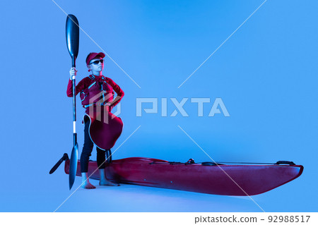 Beginner kayaker in red canoe, kayak with a life vest and a paddle isolated on blue background in neon. Concept of sport, travel, active lifestyle Beginner kayaker in red canoe, kayak with a life vest and a paddle isolated on blue background in neon. Concept of sport, travel, active lifestyle 92988517