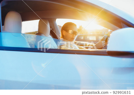 Portrait of fashion cyberpunk girl driving a white supercar in sunsert light. Woman with futuristic eyeglasses sits by the car steering wheel. Future technology concept. Selective focus, copy space 92991631