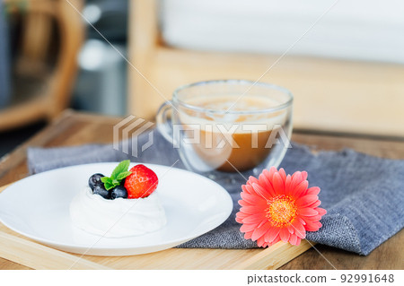 Pavlova dessert mini cake, cup of latte coffee and fresh gerbera flower on the wooden tray with gray napkin on the table. Coffee time. Breakfast for loved one. Take a break and relax. Selective focus. 92991648
