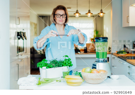 Blogger creating content for social media. Woman making photo, video with just made glass of detox green smoothie and it's ingredients on the kitchen table. Healthy dieting, cooking, weight loss blog 92991649