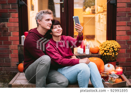 Couple taking selfies on phone during a romantic date on the porch of their home. Time at home. Autumn tea time outdoors on house entrance decorated with pumpkins, flowers and burning candles 92991895