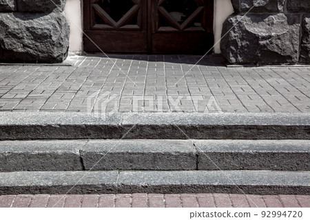 granite steps on a pedestrian pavement made of stone tiles near a building with entrance a wooden front door and a facade lined with rough stone on a sunny day, close-up, nobody. 92994720