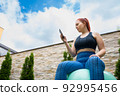 Attractive young fitness girl looking at her smart phone sitting on a pilates ball outdoors. girl exercising while sharing it on her social networks. health and wellness concept. 92995456