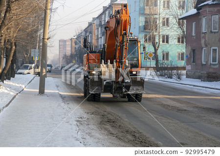 Orange excavator moving along the city winter road. Orange excavator moving along the city winter road. 92995479