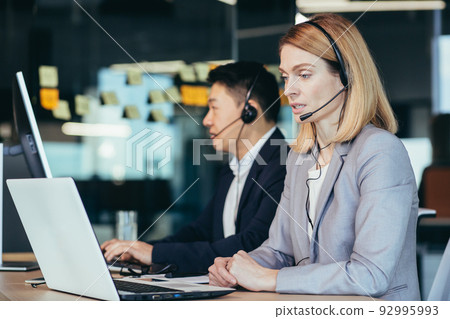 Call center, a team of workers working on a laptop and computer for a video call, a woman uses a headset, advises customers 92995993