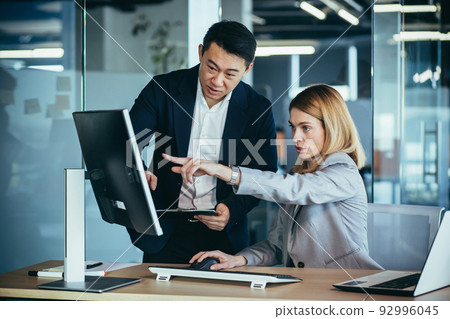 Two Asian male and female colleagues in a modern office, a woman shows the work done on the monitor, consults and discusses Two Asian male and female colleagues in a modern office, a woman shows the work done on the monitor, consults and discusses 92996045