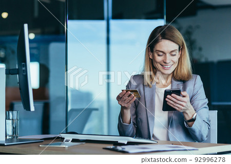 Happy and successful business woman shopping online, working in a modern office, an employee uses a phone for shopping and an application, holding a credit card Happy and successful business woman shopping online, working in a modern office, an employee uses a phone for shopping and an application, holding a credit card 92996278
