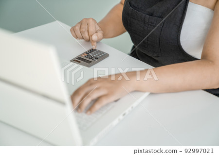 Young woman is sitting at her laptop and doing calculations on calculator. Hands, calculator, and laptop close-up. Concept of financial and project calculations and accounting. Young woman is sitting at her laptop and doing calculations on calculator. Hands, calculator, and laptop close-up. Concept of financial and project calculations and accounting. 92997021