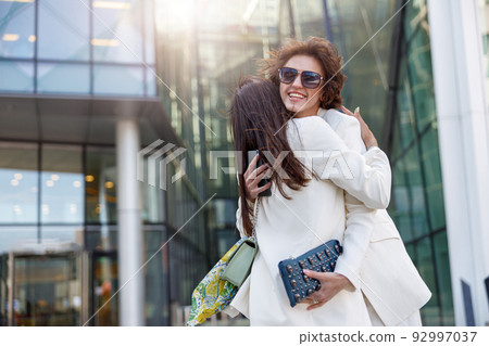 Two emotional happy woman friends in trendy outfits hugging each other on city street Two emotional happy woman friends in trendy outfits hugging each other on city street 92997037