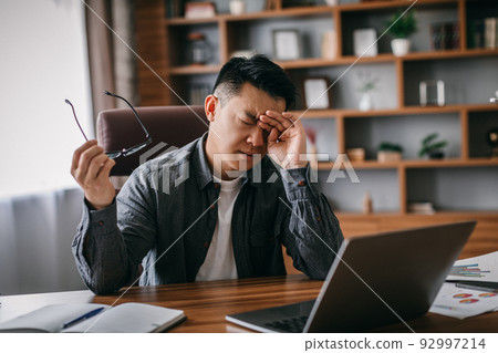 Sad tired adult chinese man takes off his glasses, rubs eyes and rests from work in home office interior 92997214