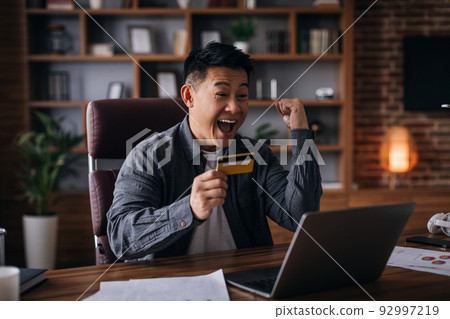 Glad adult asian man with open mouth, scream, hold credit card, looks at laptop and makes victory gesture Glad adult asian man with open mouth, scream, hold credit card, looks at laptop and makes victory gesture 92997219
