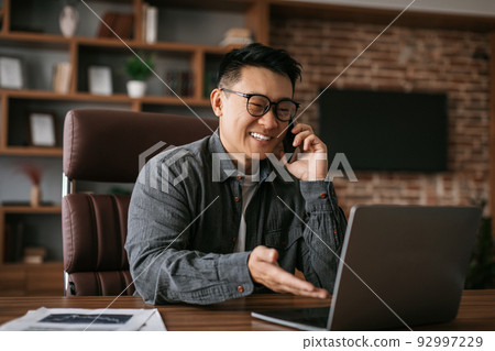 Cheerful mature japanese man in glasses calls by phone, works with laptop at table in office interior Cheerful mature japanese man in glasses calls by phone, works with laptop at table in office interior 92997229