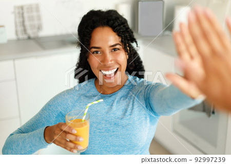 Happy african american fit lady drinking fresh juice and giving high five to friend, standing in kitchen interior Happy african american fit lady drinking fresh juice and giving high five to friend, standing in kitchen interior 92997239