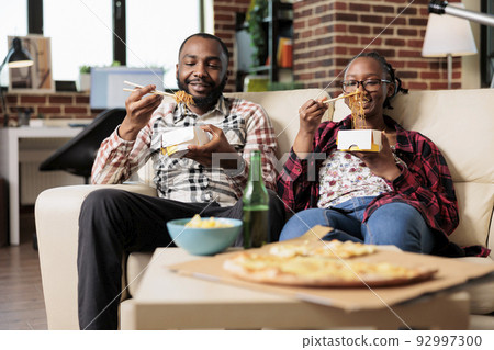 Man and woman eating noodles from takeout delivery box, having fun with fast food takeaway meal on couch. Using chopsticks to eat dinner and watching movie or film on television program. 92997300