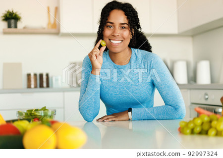 Excited fit african american lady eating fruits for breakfast, holding grape and smiling at camera, sitting in kitchen Excited fit african american lady eating fruits for breakfast, holding grape and smiling at camera, sitting in kitchen 92997324