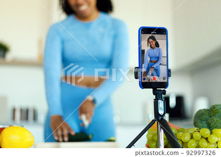 Slim african american woman in sportswear preparing fresh salad and shooting video on smartphone camera in kitchen Slim african american woman in sportswear preparing fresh salad and shooting video on smartphone camera in kitchen 92997326