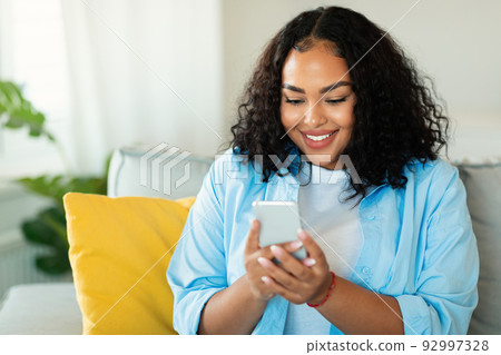 Cheerful African American Woman Using Smartphone Sitting At Home Cheerful African American Woman Using Smartphone Sitting At Home 92997328
